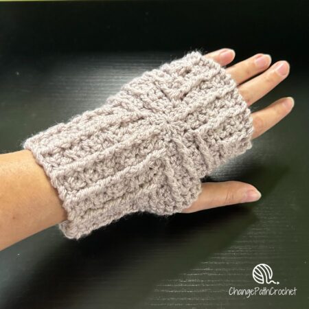 A left hand in a crocheted fingerless glove. The mitt has a cable pattern at right angles from the center of the back of the hand.