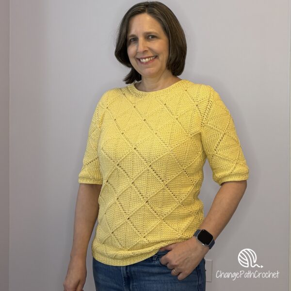 Woman standing with one hand in her pocket, wearing a yellow, handmade Tunisian crochet shirt with half sleeves.