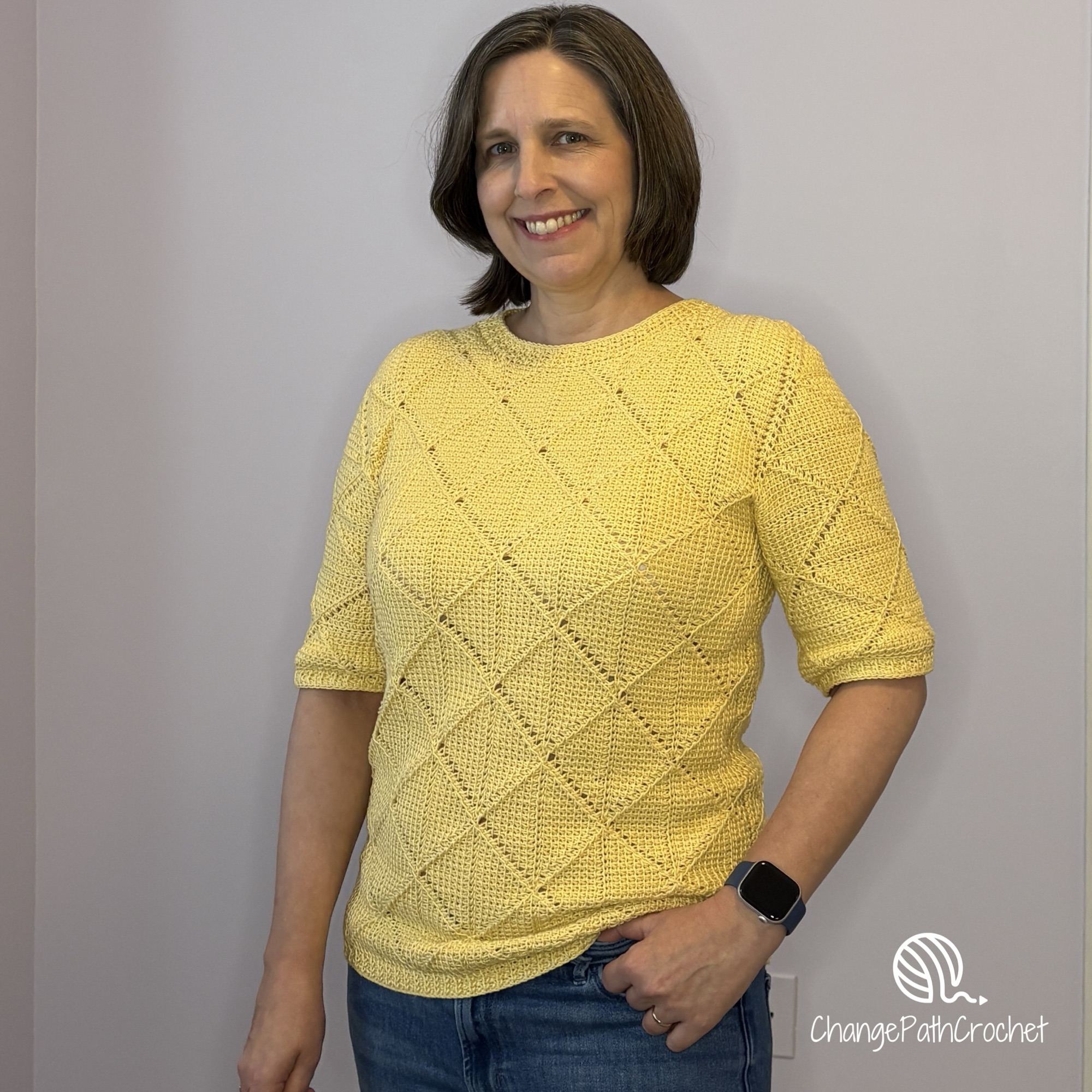 Woman standing with one hand in her pocket, wearing a yellow, handmade Tunisian crochet shirt with half sleeves.