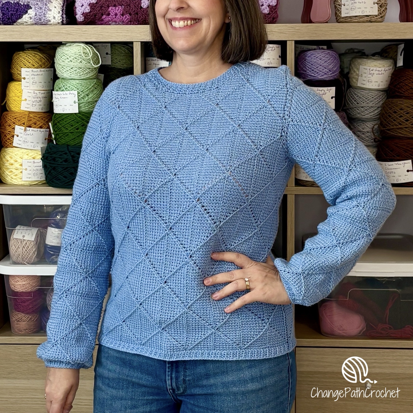Woman standing with hand on hip, wearing a blue long-sleeve sweater with a diamond motif.